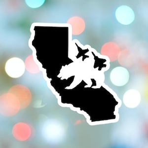 Può includere: Un adesivo in bianco e nero con il contorno della California. All'interno del contorno ci sono un orso bianco e due jet da combattimento. L'adesivo ha un bordo bianco ed è posto su uno sfondo sfocato.