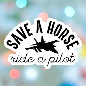 May include: A white sticker with the text "SAVE A HORSE ride a pilot" in a semi-circular design. A black silhouette of a jet fighter is in the center. The background is a blurred mix of colors.