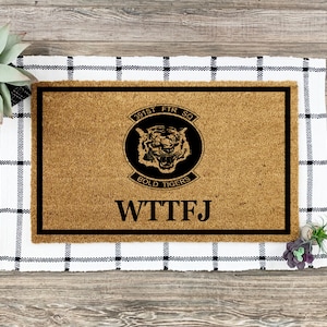 Può includere: Un tappetino da porta marrone con bordo nero e il testo "391ST FTR SQ BOLD TIGERS" e "WTTFJ" in nero.