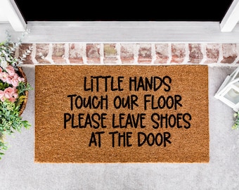 May include: A rectangular doormat made of natural brown coir fiber. The mat features black text that reads "LITTLE HANDS TOUCH OUR FLOOR PLEASE LEAVE SHOES AT THE DOOR". The mat is placed on a concrete surface near a doorway.