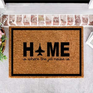 May include: A brown coir doormat with black trim and the text "HOME is where the jet noise is" with a black silhouette of a jet plane in place of the "O".