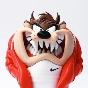 Peut inclure: Figurine détaillée du Diable de Tasmanie, également connu sous le nom de Taz, avec un large sourire révélant des dents pointues. La figurine porte une veste rouge sur une chemise blanche avec un logo Nike noir. Les traits du visage et la fourrure brune du personnage sont bien définis.