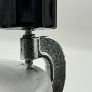 Könnte beinhalten: Nahaufnahme eines schwarzen, strukturierten Knopfes, der an einer silberfarbenen Klemme befestigt ist. Die Klemme hat einen gebogenen Griff und einen Gewindeschraubenmechanismus. Der Hintergrund ist schlicht weiß.