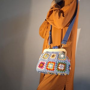 May include: A blue crochet purse with a colorful granny square design. The purse has a light-colored frame and a blue shoulder strap. The bag is a handmade item.