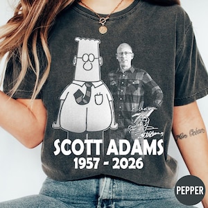 May include: Charcoal grey t-shirt with a cartoon figure and a portrait of a man. The shirt displays the text "SCOTT ADAMS 1957 - 2026" in white. The design includes a signature and the name "Michelle".