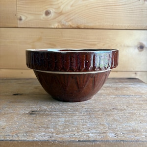 May include: A brown ceramic mixing bowl with a ribbed design around the rim and a textured exterior. The bowl is set on a wooden surface, showcasing its vintage aesthetic. This kitchenware item is ideal for baking and food preparation.