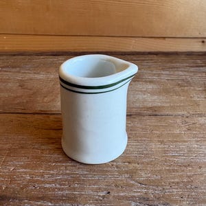 May include: A small white ceramic creamer pitcher featuring a simple design with two thin, dark green horizontal stripes encircling the upper rim. The pitcher has a smooth, cylindrical body and a small, integrated pouring spout on one side.