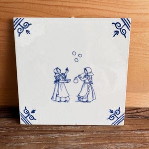 May include: Square white ceramic tile with a blue and white illustration. The image depicts two figures in traditional attire, with one appearing to juggle three objects. Decorative blue corner accents complete the design.