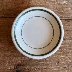 May include: A small, round, white ceramic bowl featuring a simple dark green band encircling the inner rim and a thinner dark green circular line centered on the interior base. The bowl has a smooth, glossy finish and a clean, circular design.