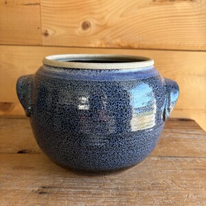 May include: A round, blue ceramic pot with two handles. The pot has a speckled texture and a light-colored rim. The background is a wooden surface and a wooden wall.