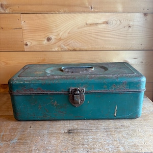 May include: A vintage teal metal toolbox with a hinged lid and a rusty metal clasp. The rectangular box has a handle on top and shows signs of wear and age, with visible rust and scratches. The toolbox sits on a wooden surface.