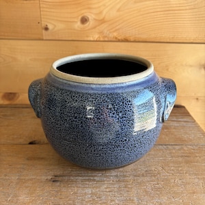 May include: A speckled blue ceramic pot with two handles. The pot has a wide opening and a dark interior. The exterior has a textured, speckled pattern in shades of blue and gray. The pot is sitting on a wooden surface.