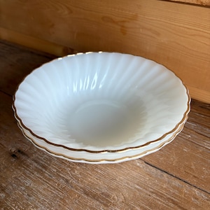 May include: Two white, vintage-style bowls with a gold trim. The bowls have a fluted design and are stacked on top of each other. The bowls are on a wooden surface.