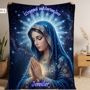 May include: A blue blanket featuring an illustration of a woman in a blue veil with hands clasped in prayer. The blanket has the text "Wrapped in Marian love" and the name "Jennifer". The background is a starry night.