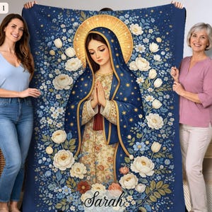 May include: A navy blue blanket featuring a religious illustration of a woman with a gold halo, surrounded by white and blue flowers. The blanket has the text "Sarah" at the bottom. The blanket is held by two people.