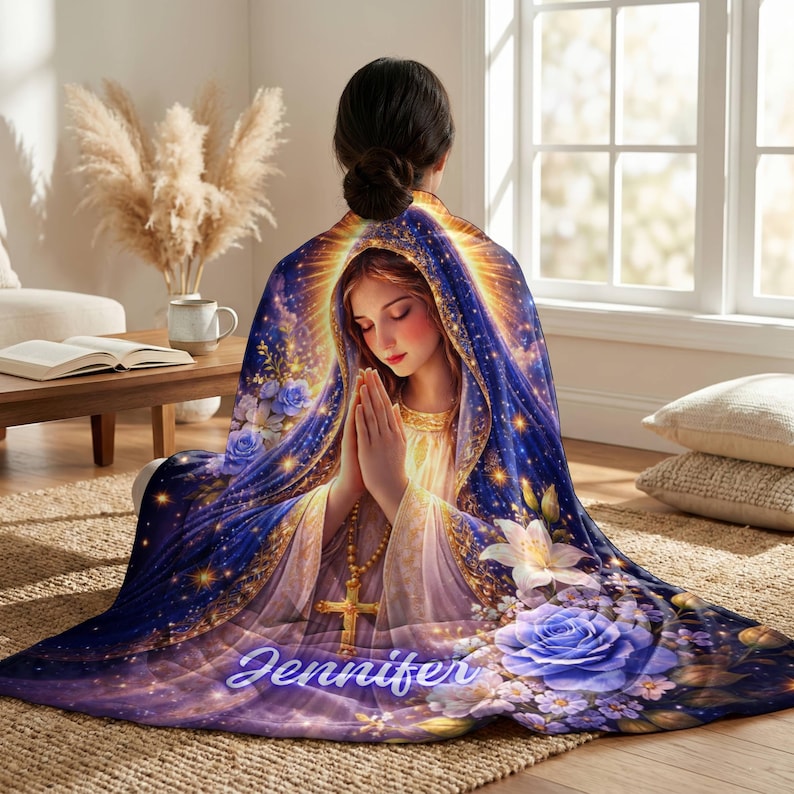 May include: A plush blanket featuring a religious image of a woman in prayer, draped in a blue and gold cloak. The design includes floral accents, a cross pendant, and the name "Jennifer" in script. The blanket is on a woven rug.