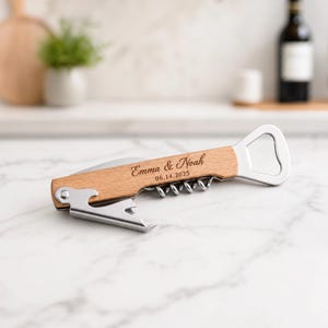 Peut inclure: Un tire-bouchon multifonction doté d'un manche en bois clair gravé avec le texte Emma & Noah et la date 06.14.2025. L'outil comprend un tire-bouchon en acier inoxydable, une lame coupe-capsule et un décapsuleur intégré au design.