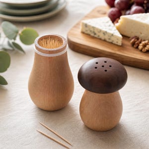 Peut inclure: Un porte-cure-dents en bois clair avec un bord blanc et une salière assortie en forme de champignon, dotée d'un capuchon marron foncé percé de cinq petits trous. Deux cure-dents en bois sont posés devant ces deux objets de cuisine en bois.