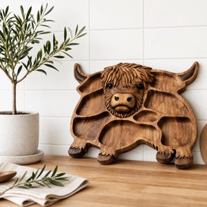 May include: A brown wooden serving tray shaped like a Highland cow, featuring a carved three-dimensional head in the center and multiple recessed compartments for food items, with visible wood grain and dark stained details on the horns and hooves.