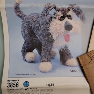 May include: A Butterick 3856 sewing pattern for a Jim Henson's Fraggle Rock Sprocket plush toy. The paper envelope features a photograph of a shaggy black and white dog puppet with a pink tongue, alongside printed instructions and material lists.