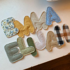 May include: Handmade fabric letters spelling out the names "EMMA" and "ELLA" and "JAH". The letters are in various colors and patterns, including floral, polka dots, stripes, and plaid. Each letter has a scalloped edge.