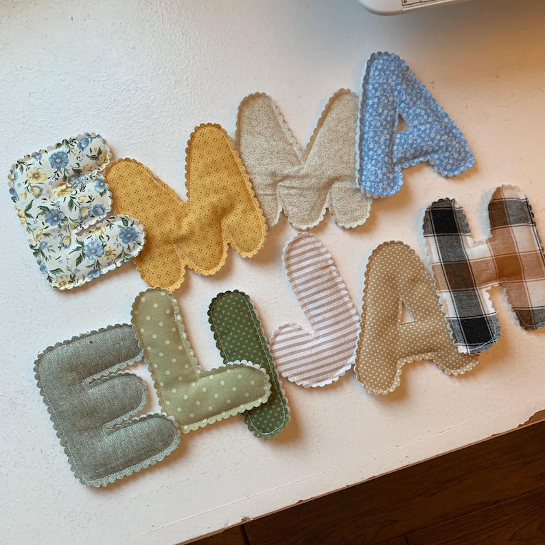 May include: Handmade fabric letters spelling out the names "EMMA" and "ELLA" and "JAH". The letters are in various colors and patterns, including floral, polka dots, stripes, and plaid. Each letter has a scalloped edge.