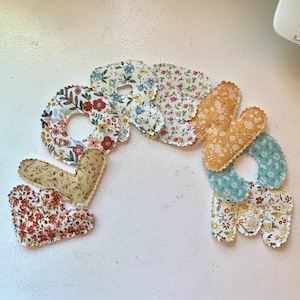 May include: A handmade fabric word "Love" in a variety of floral patterns. The letters are in a mix of colors, including red, blue, yellow, and green, with scalloped edges. The word is arranged in a curved shape.