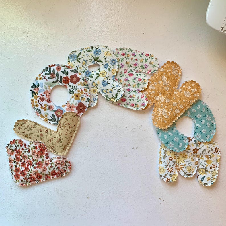 May include: A handmade fabric word "Love" in a variety of floral patterns. The letters are in a mix of colors, including red, blue, yellow, and green, with scalloped edges. The word is arranged in a curved shape.