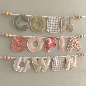 May include: Fabric name banners spelling out the names "COEN", "SOFIA", and "OWEN". Each letter is a different fabric pattern, including polka dots, gingham, stripes, and floral prints. The letters are strung on white cord with wooden beads.