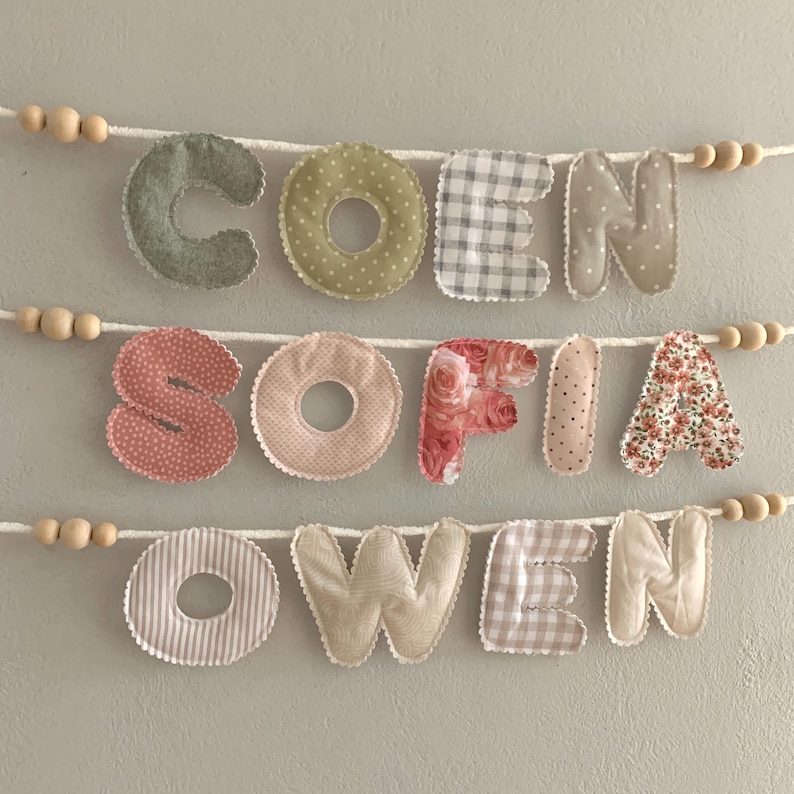 May include: Fabric name banners spelling out the names "COEN", "SOFIA", and "OWEN". Each letter is a different fabric pattern, including polka dots, gingham, stripes, and floral prints. The letters are strung on white cord with wooden beads.