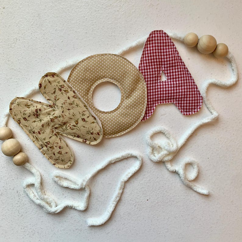May include: A decorative banner spelling "NOA" with fabric letters. The "N" is floral, the "O" is tan with white polka dots, and the "A" is red and white gingham. Wooden beads and white fluffy cord complete the design.