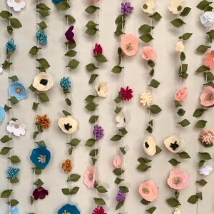 May include: A garland of felt flowers and leaves in various colors, including pink, blue, white, green, and brown. The garland is strung on a white string and is perfect for adding a touch of whimsy to any room.