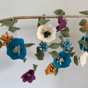 May include: A felt garland with teal, white, and purple flowers and green leaves. The garland is strung on a branch.