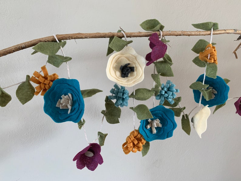May include: A felt garland with teal, white, and purple flowers and green leaves. The garland is strung on a branch.