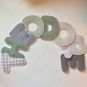 May include: A handmade fabric banner spelling "GODDAMA" in various patterns and colors. The letters are in shades of green, gray, and beige, with polka dots, stripes, and plaid designs. The banner is likely for nursery or home decor.