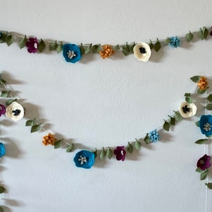 May include: Two felt flower garlands with green leaves, blue, white, and purple flowers. The garlands are hanging on a white wall.