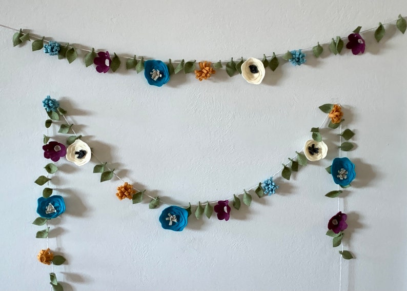 May include: Two felt flower garlands with green leaves, blue, white, and purple flowers. The garlands are hanging on a white wall.