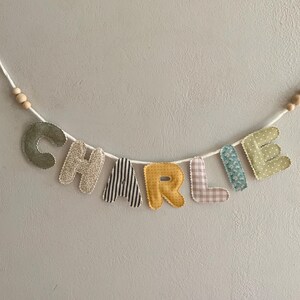 May include: A handmade banner spelling "CHARLIE" in colorful fabric letters. The letters are attached to a white string with wooden beads. Each letter has a unique pattern, including stripes, polka dots, and floral designs.