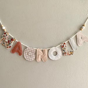 May include: A fabric banner spelling "MAGNOLIA" in various patterned letters. The letters are sewn onto a white cord, with wooden beads at each end. The letters feature floral, gingham, and striped patterns in shades of red, pink, and beige.