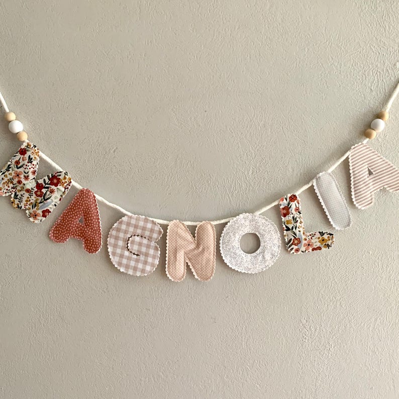 May include: A fabric banner spelling "MAGNOLIA" in various patterned letters. The letters are sewn onto a white cord, with wooden beads at each end. The letters feature floral, gingham, and striped patterns in shades of red, pink, and beige.