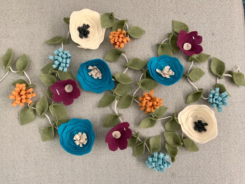 May include: A felt garland with teal, white, burgundy, and orange flowers. The garland is strung with green felt leaves.