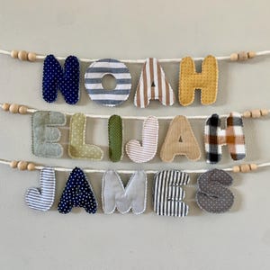 May include: A handmade fabric banner spelling out the names "NOAH ELIJAH JAMES" in various patterned letters. The letters are in blue polka dots, stripes, and plaid, and are strung on a white cord with wooden beads.