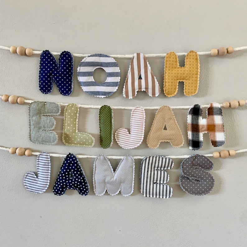 May include: A handmade fabric banner spelling out the names "NOAH ELIJAH JAMES" in various patterned letters. The letters are in blue polka dots, stripes, and plaid, and are strung on a white cord with wooden beads.