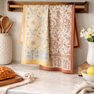 May include: Two rectangular kitchen towels hanging on a wooden rack. The left towel features a yellow and blue floral pattern with a solid yellow border. The right towel displays a rust-colored intricate botanical print with a matching rust border.
