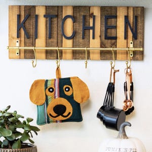 May include: Wooden kitchen sign with the word "KITCHEN" in black letters. A brass-colored rod with hooks hangs below the sign, holding a dog-shaped pot holder and measuring cups. The pot holder is green, orange, and striped.