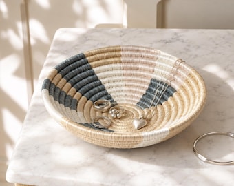 May include: A shallow, round, coiled basket featuring a woven pattern of natural tan, cream, and dark charcoal gray fibers. Inside the basket are three silver-toned rings and a silver heart-shaped pendant on a delicate chain, with a silver bangle nearby.