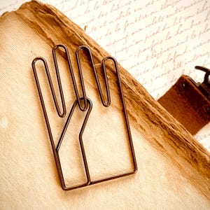 May include: A wire bookmark shaped like two hands holding each other. The bookmark is made of brown wire and is resting on a page of a book.