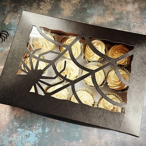 May include: A black box with a spiderweb cutout design. The box is filled with cupcakes with white frosting.