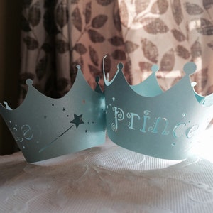 Puede incluir: Dos coronas de papel azul claro con un dise&ntilde;o de estrella y varita en una corona y la palabra "Prince" en la otra corona.