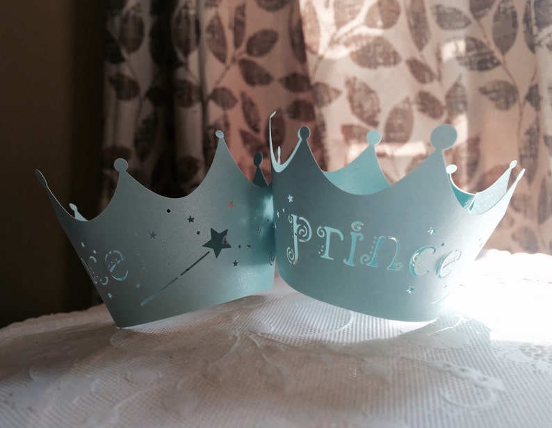 Puede incluir: Dos coronas de papel azul claro con un dise&ntilde;o de estrella y varita en una corona y la palabra "Prince" en la otra corona.